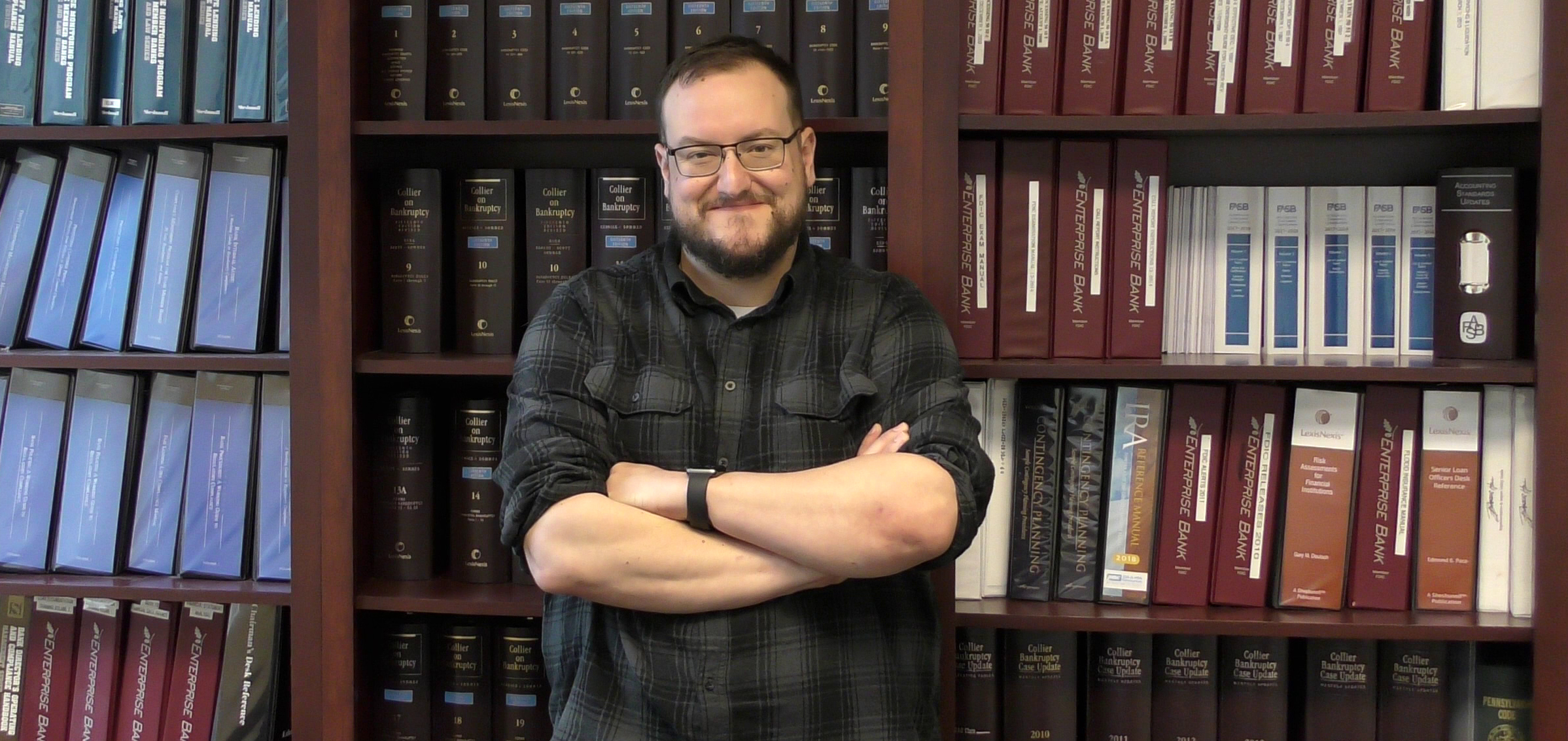 A smiling gentleman stands with his arms crossed. He is wearing a flannel shirt, glasses, and he has a beard. He stands in front of a bookcase.